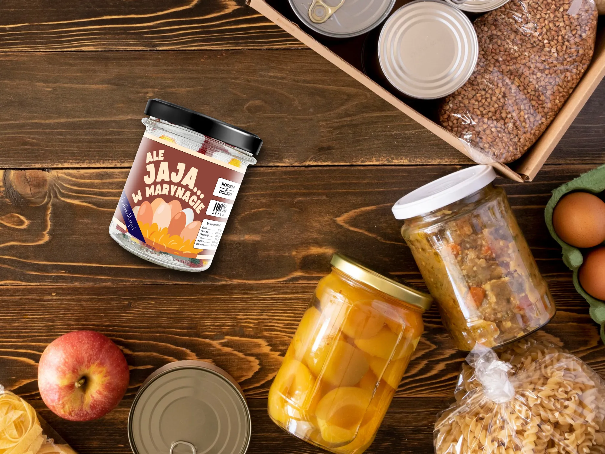 Różnorodne produkty spożywcze rozłożone na drewnianym stole, a wśród nich wyróżnia się słoik z zabawną etykietą „Ale Jaja w Marynacie”, zawierający kreatywnie zapakowane skarpetki prezentowe. Obok niego znajdują się słoiki z przetworami, puszki, jabłka, jajka i makaron, a także drewniana skrzynka z zapasami.
